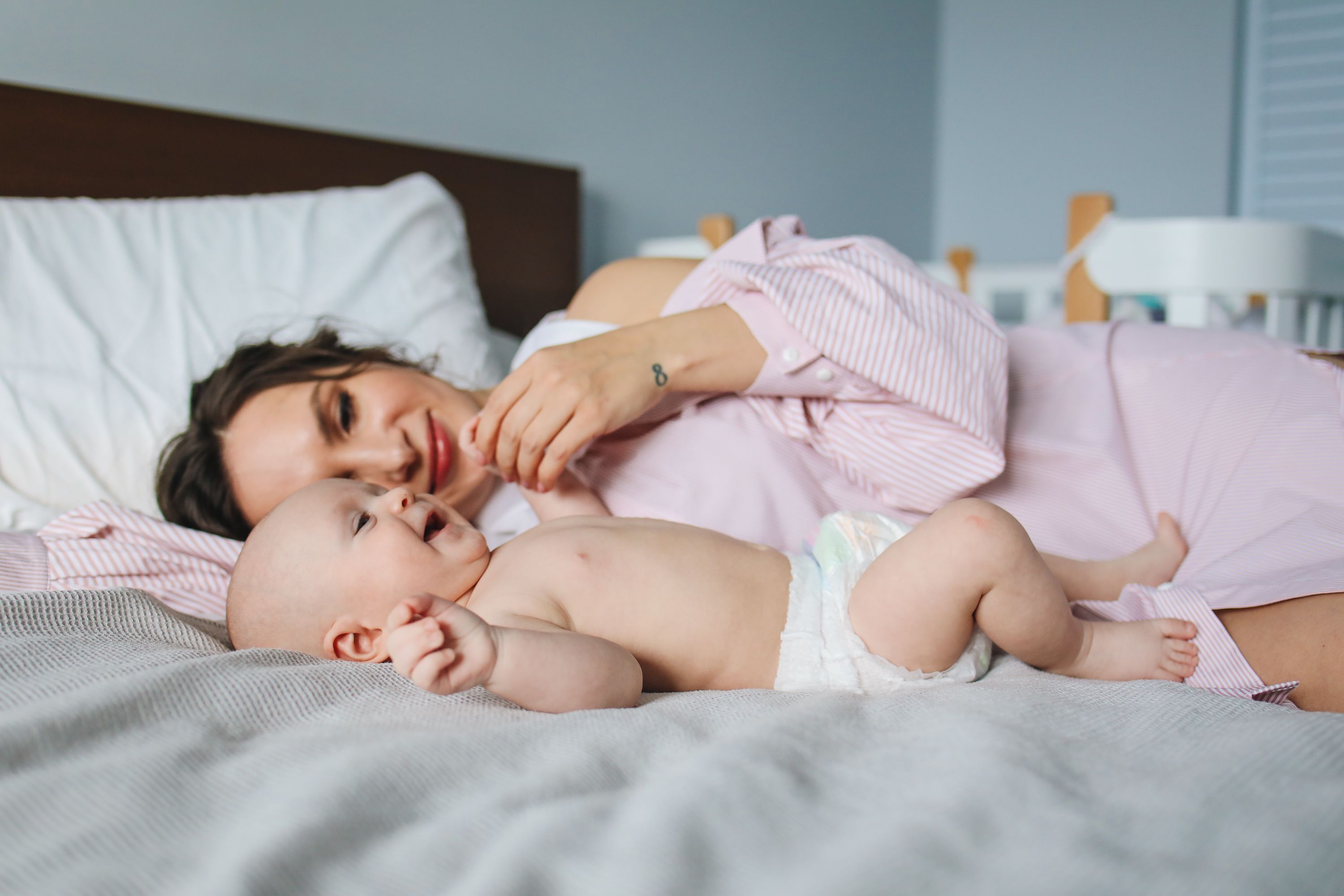 Mãe deitada de lado na cama, olhando para o bebê recém-nascido deitado ao lado dela, em um quarto claro